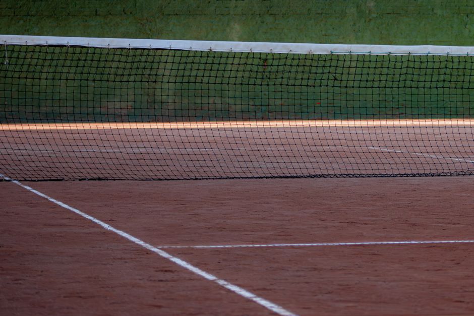 A close-up view of a tennis court with a black net stretched across the reddish-brown clay surface, which is marked with white boundary lines. The net is positioned at the center of the image, dividin