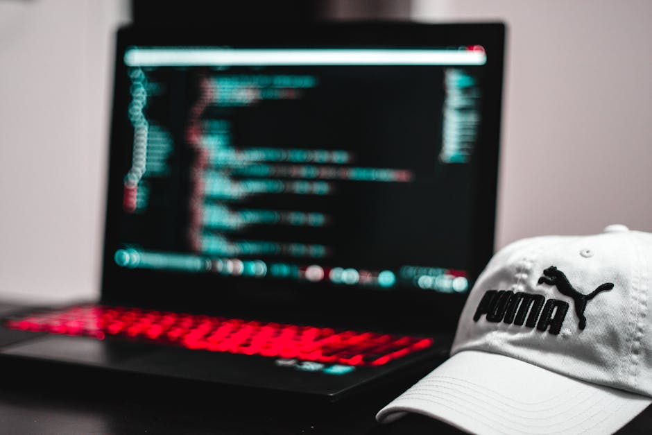 Close-up of a person's hands operating a laptop computer placed on a dark surface, with the screen displaying lines of coding or script in a dark theme interface. The background is blurred, suggesting