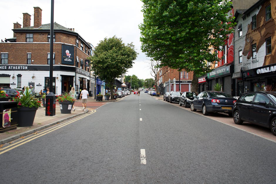 A residential street scene during daytime showing a narrow road with parked cars lining both sides, including a silver Audi and a white vehicle, with a red double-decker bus approaching in the distanc